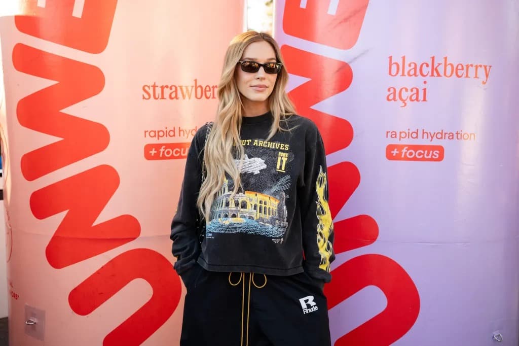 Alex Cooper wearing sunglasses and a graphic sweatshirt, standing in front of large Unwell beverage-branded pillars.
