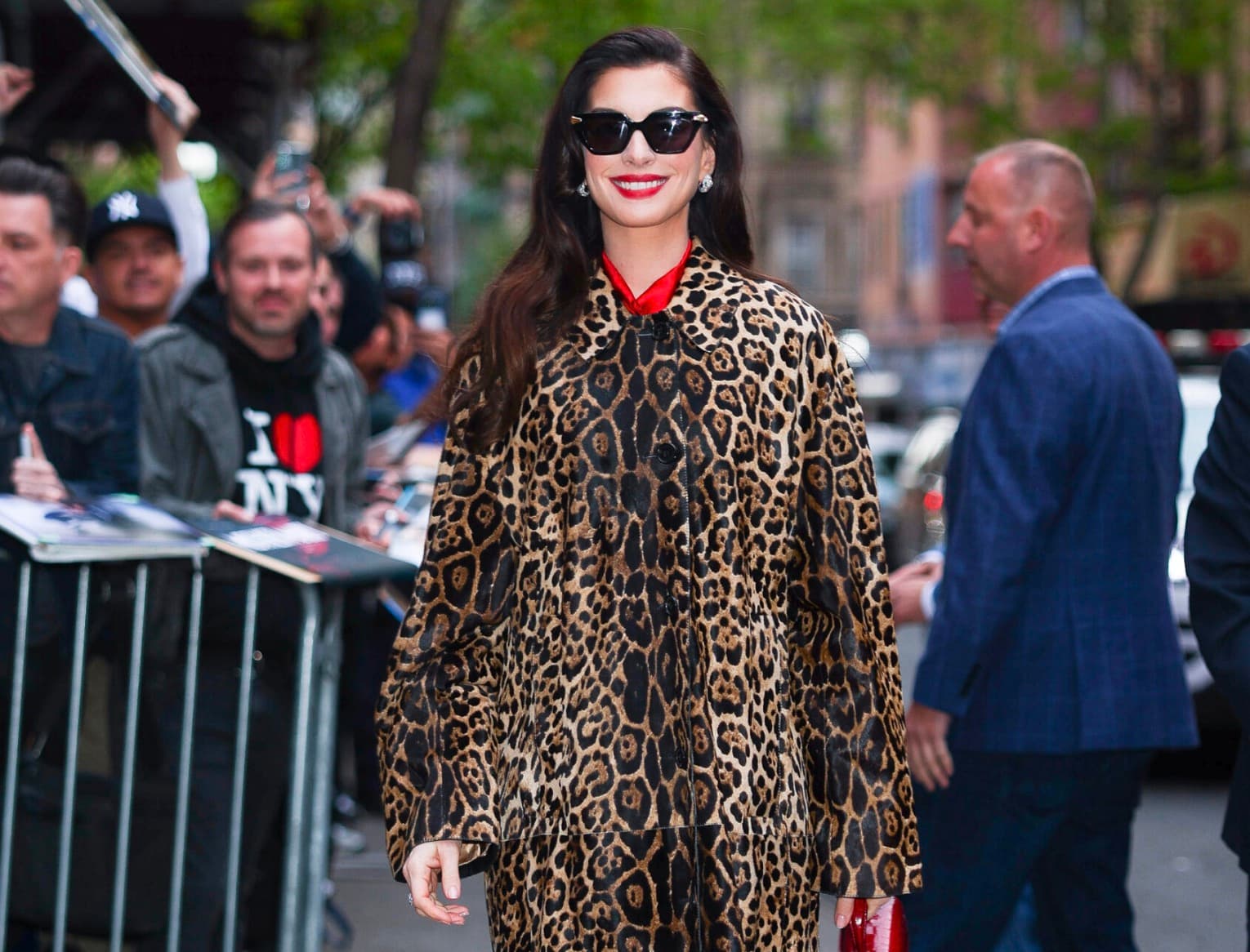 Anne Hathaway smiling in sunglasses and a leopard-print coat, greeting fans on a New York street.