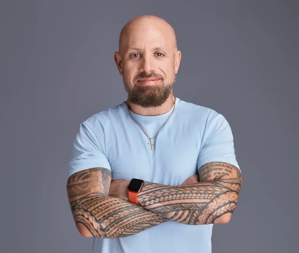 Mark McQuade smiling with arms crossed, showing tattooed forearms, against a gray background.