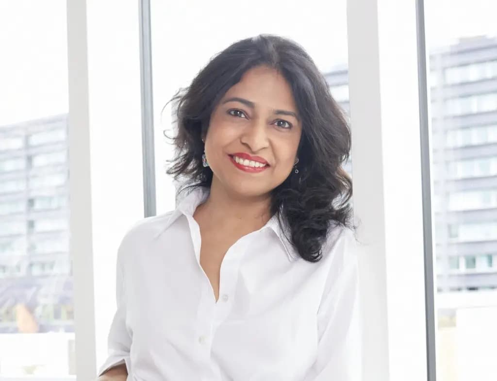 Asmita Dubey smiling in a white shirt in front of floor-to-ceiling windows overlooking a city.