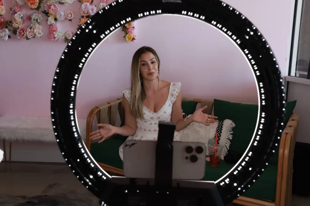A woman filming herself with a smartphone inside a ring light, seated on a couch during a content creation session.