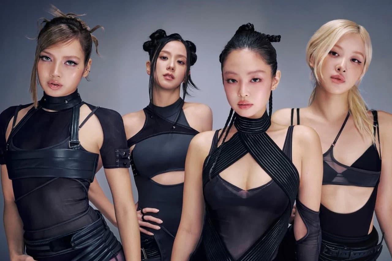 Blackpink's four members pose together in coordinated black outfits with structured straps and cutouts.
