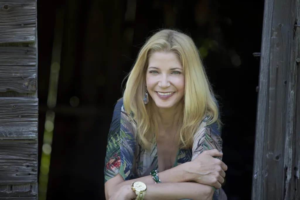 Candace Bushnell smiling in a patterned jacket, leaning in a rustic wooden doorway.
