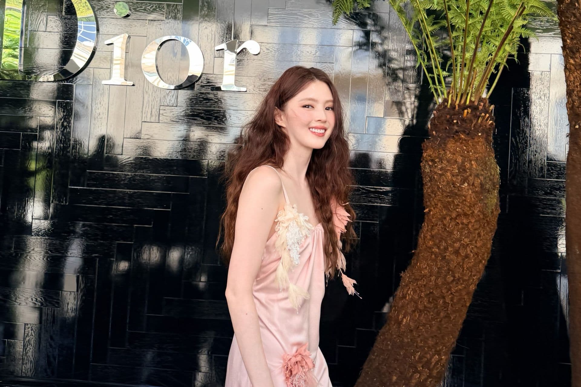 A woman in a pink feathered and floral-embellished slip dress smiles outside a Dior show backdrop.