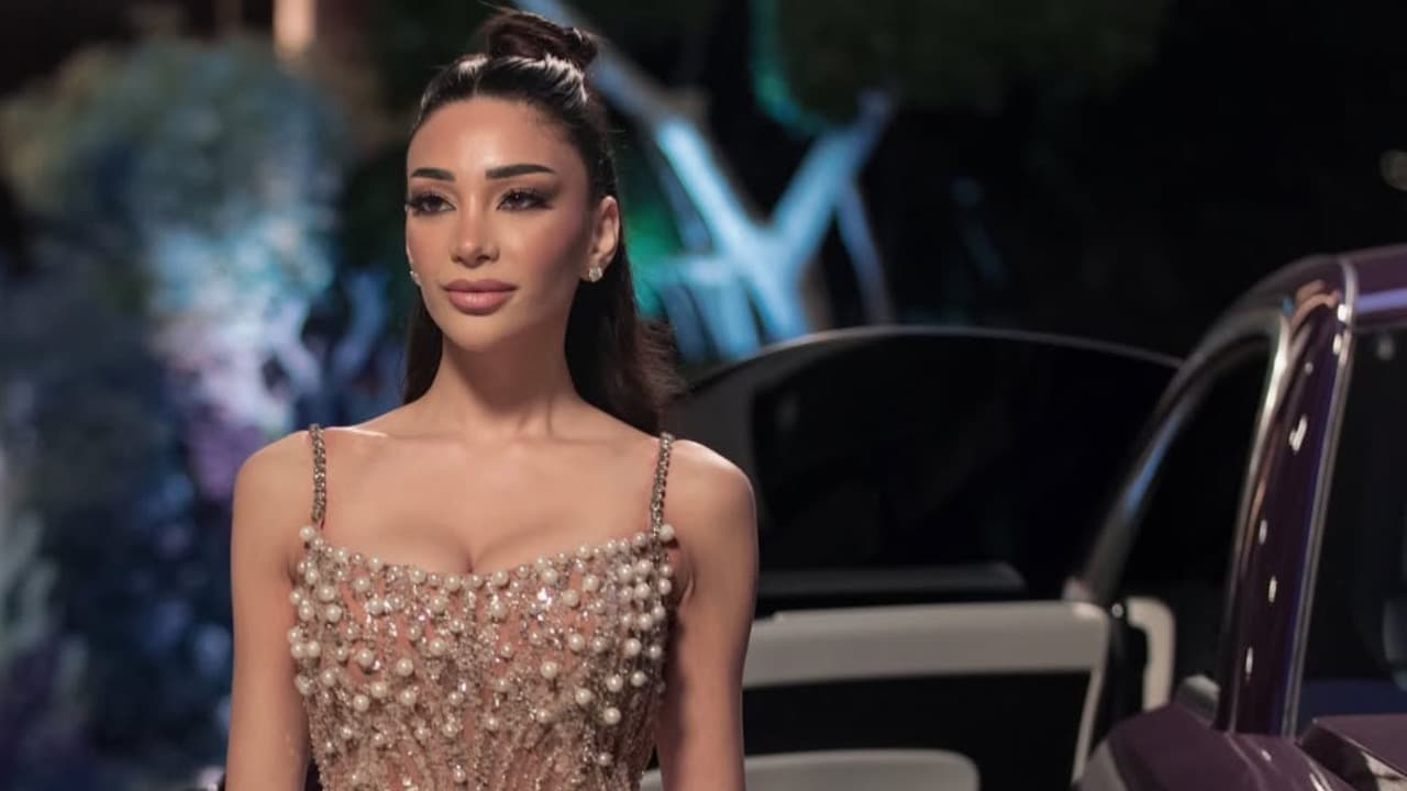 A woman in a pearl and crystal embellished gown stands beside a luxury car at night.
