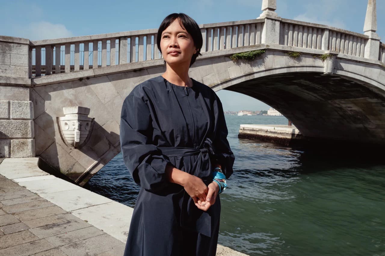Artist Dian Suci stands beside a canal bridge in Venice wearing a dark belted coat.