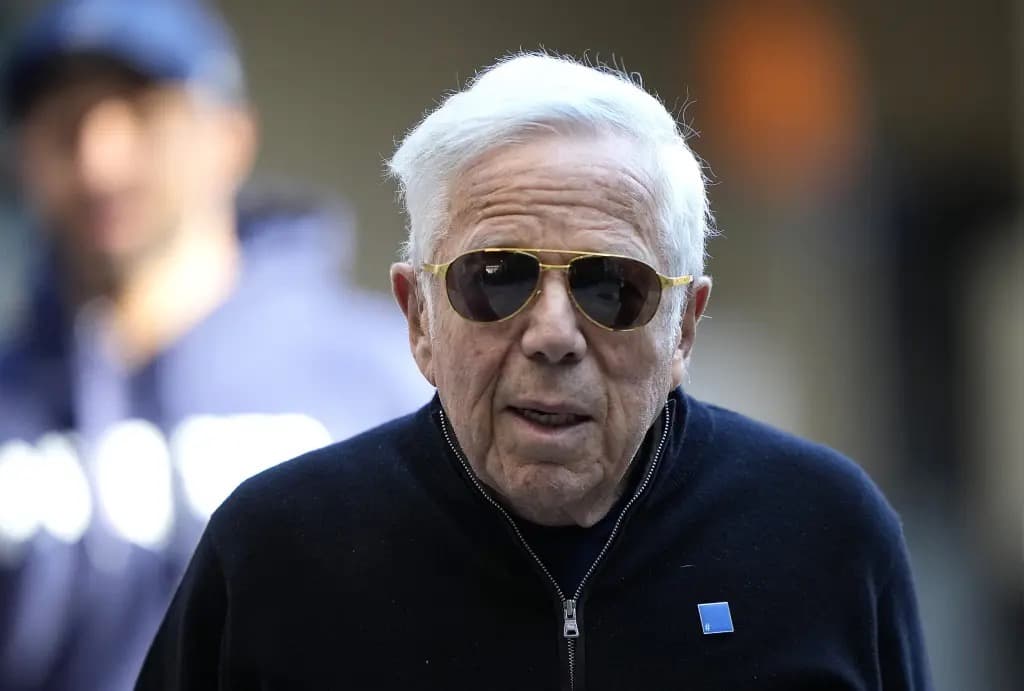 Robert Kraft wearing sunglasses and a navy zip-up sweater, walking outdoors.
