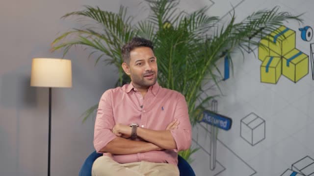 Kapil Thirani seated with arms crossed during an interview, with illustrated shipping boxes on the wall behind him.