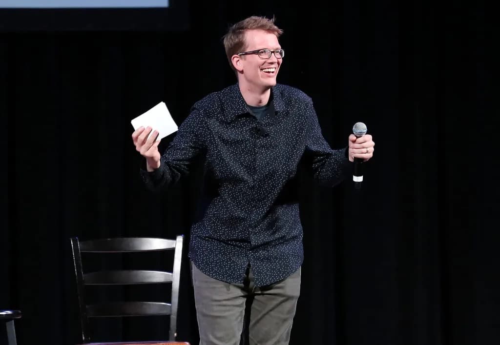 Hank Green smiling on stage while holding a microphone and an index card.