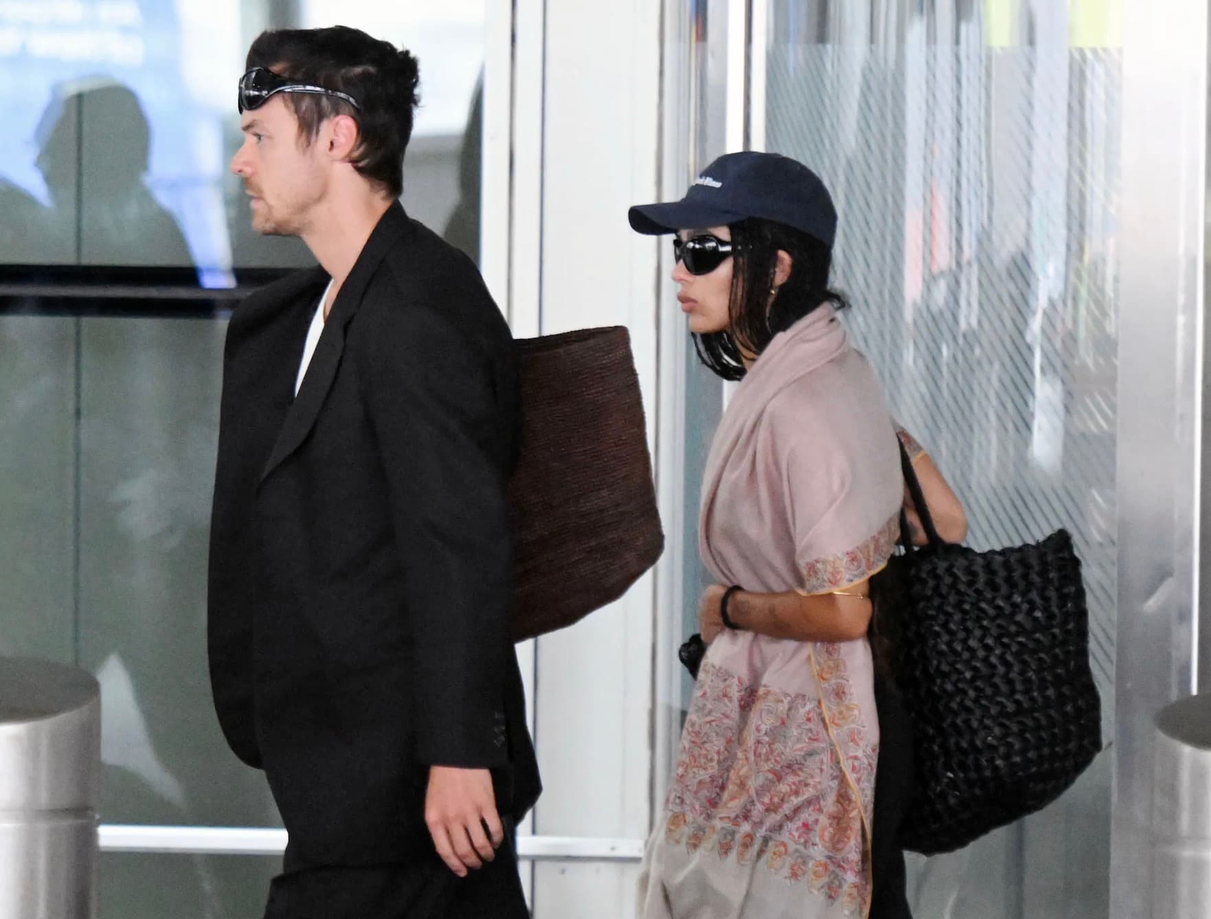 Harry Styles and Zoë Kravitz walk through an airport wearing sunglasses, Styles in black and Kravitz in a pale pink shawl.