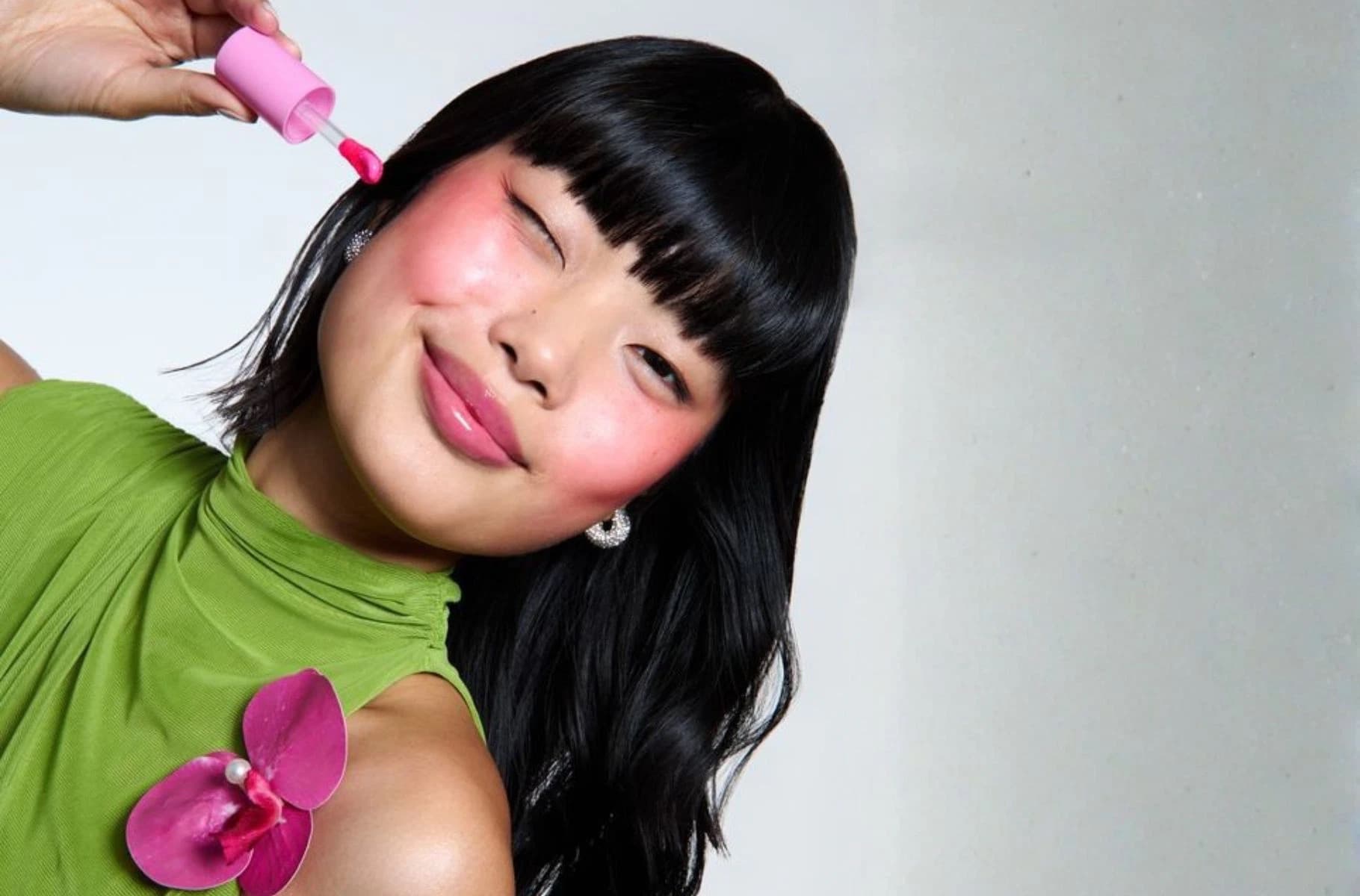 A model with a flushed pink blush application, holding a cream blush wand near her cheek.