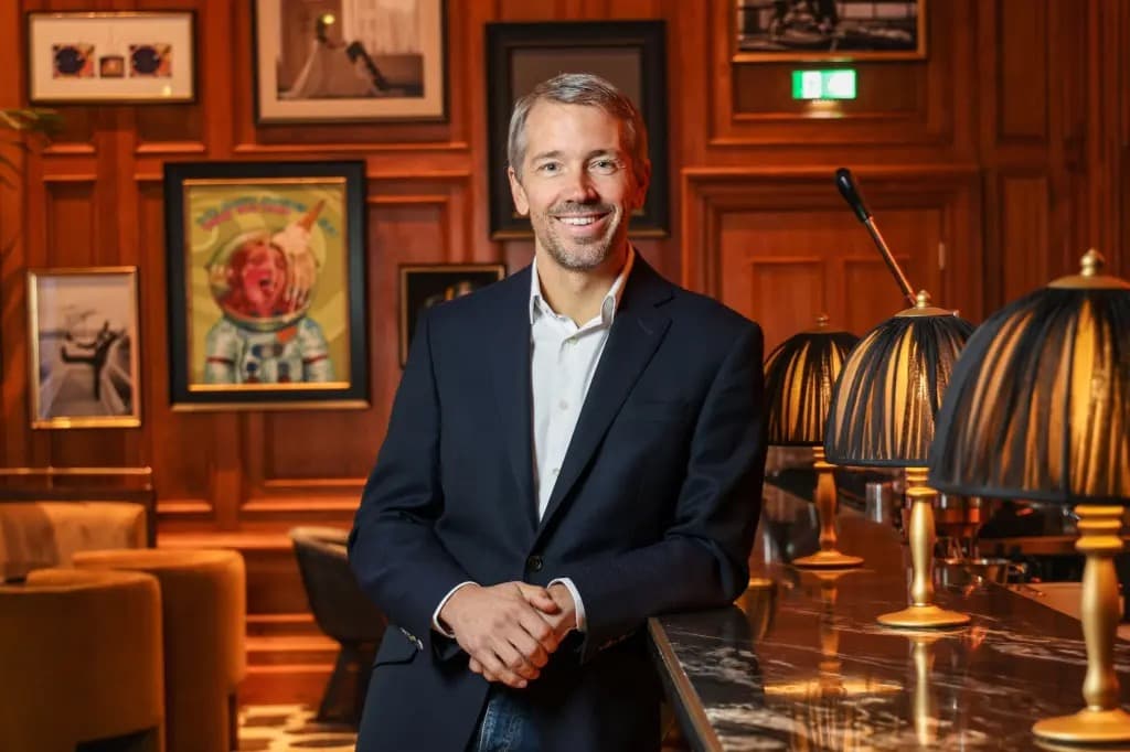 Josh Bayliss smiling in a navy blazer, leaning against a bar counter in a wood-paneled room.