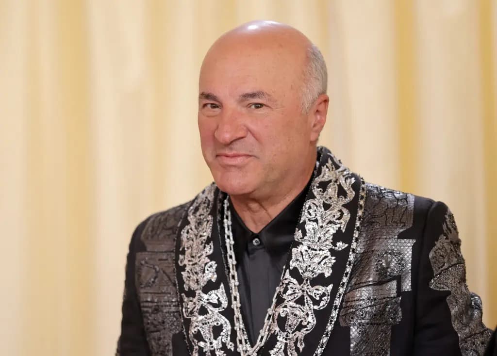 Kevin O'Leary smiling in an embellished black jacket in front of a cream curtain backdrop.