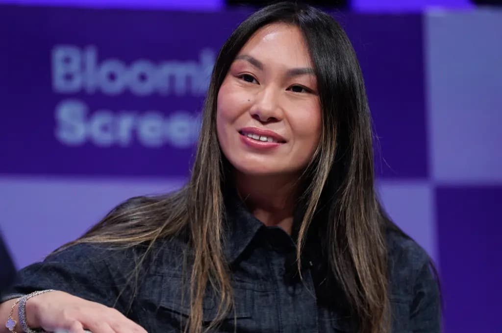 Lucy Guo smiling in a dark shirt during a Bloomberg interview, seated in front of a purple backdrop.