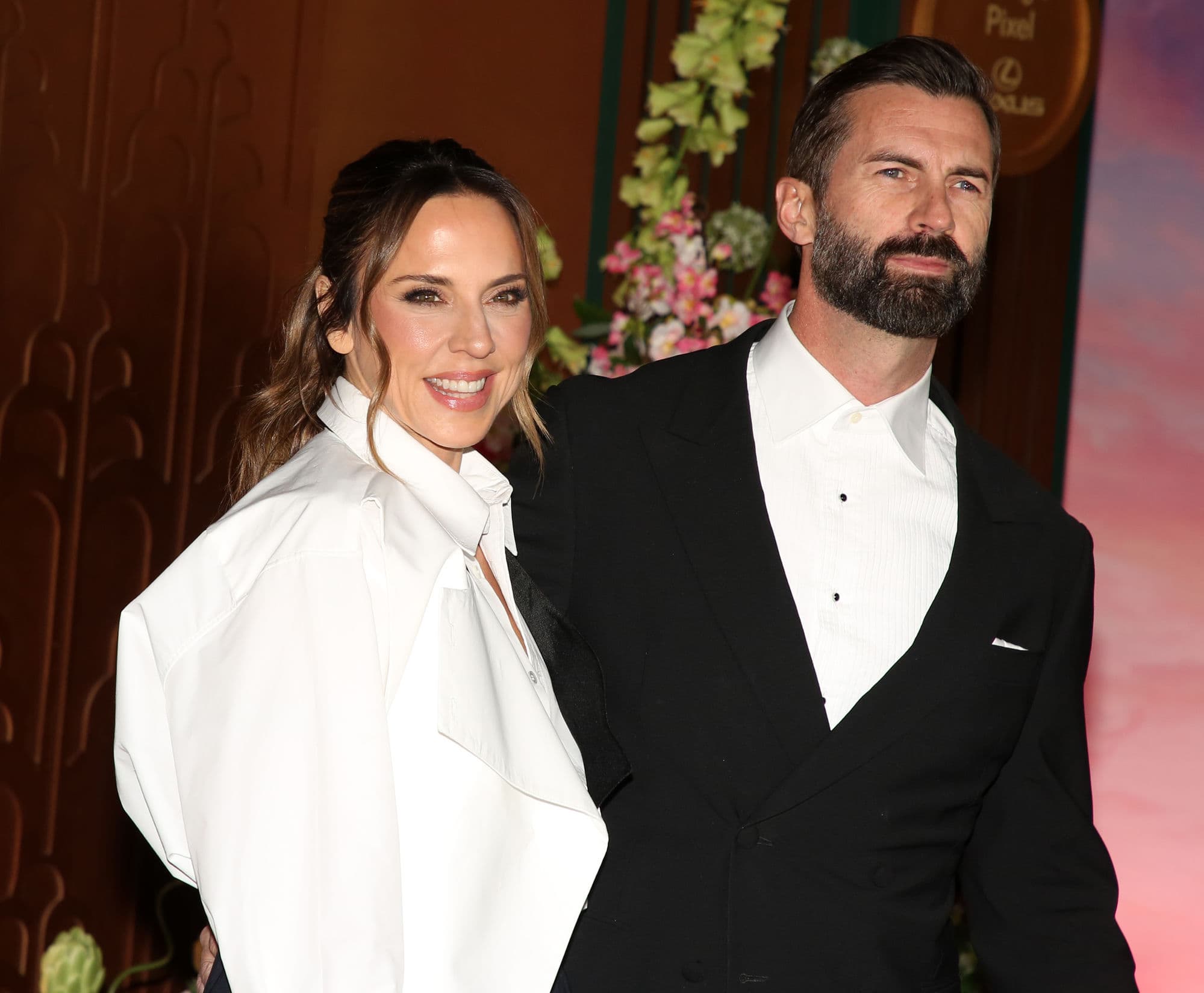 Mel C smiles beside her husband Chris Dingwall, who wears a black tuxedo, at a red carpet event.