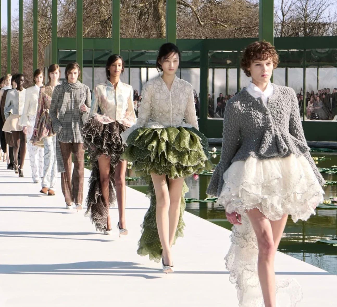 Models walk a runway built over a garden pond in ruffled, textured couture looks.