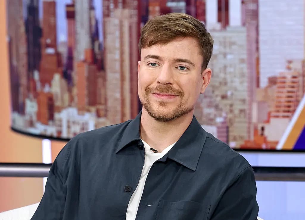 MrBeast (Jimmy Donaldson) smiling in a dark jacket during a television interview, with a city skyline backdrop.