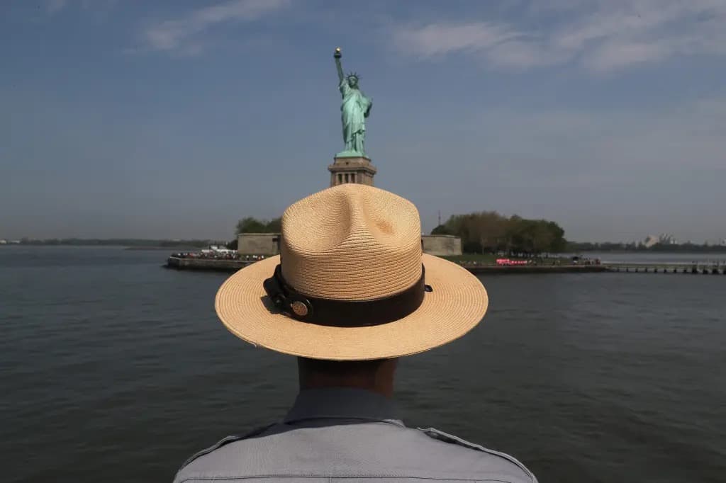 A park ranger in a wide-brimmed hat viewed from behind, facing the Statue of Liberty across the water.