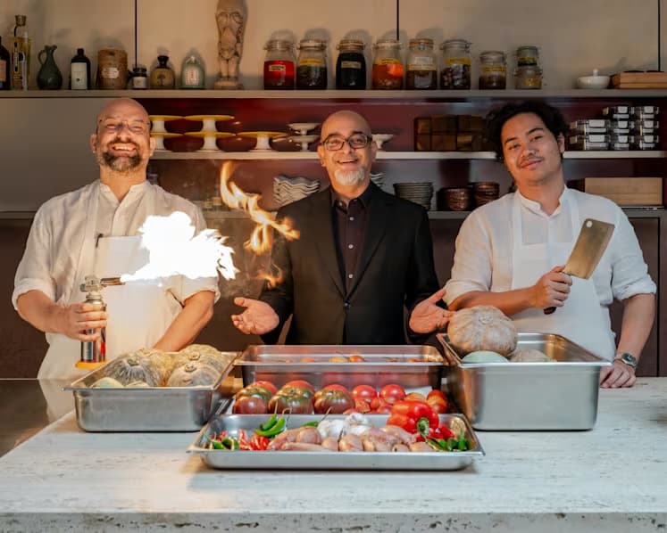 Chef Ivan Brehm, Huzir Sulaiman, and an actor from Checkpoint Theatre pose in Restaurant Nouri's open kitchen surrounded by trays of produce.