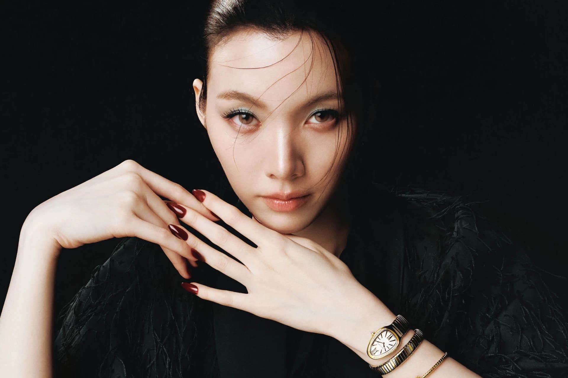 Orm Kornnaphat poses with her hand near her face wearing gold jewellery against a dark background.