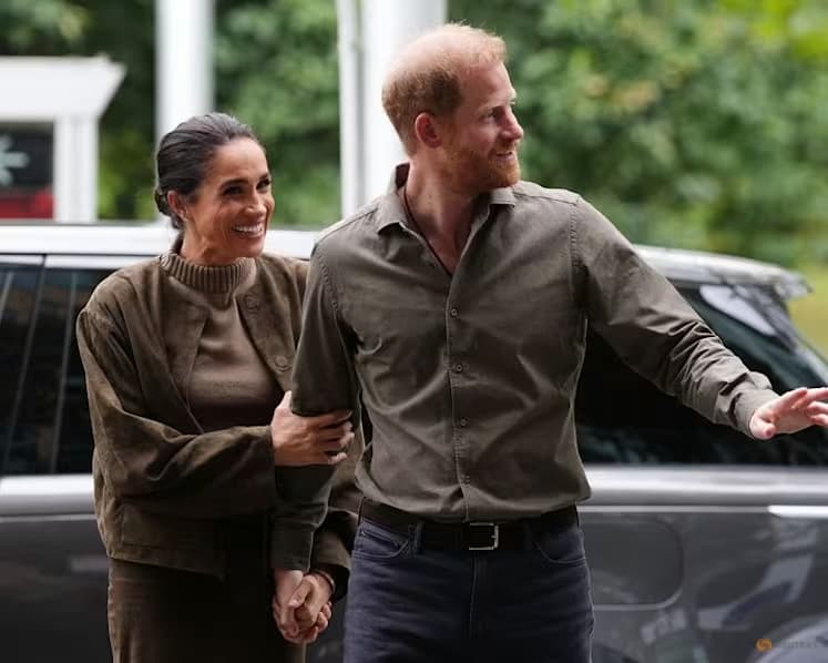 Prince Harry and Meghan Markle walk arm in arm outside, smiling, in front of a parked car.
