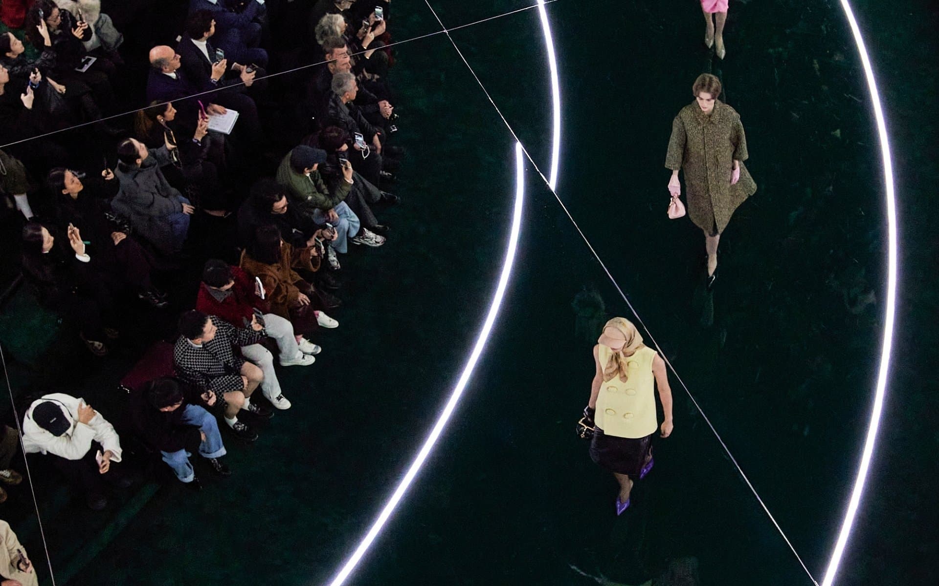 An aerial view of a Gucci runway show, with models walking between glowing curved light installations as the audience watches from the side.