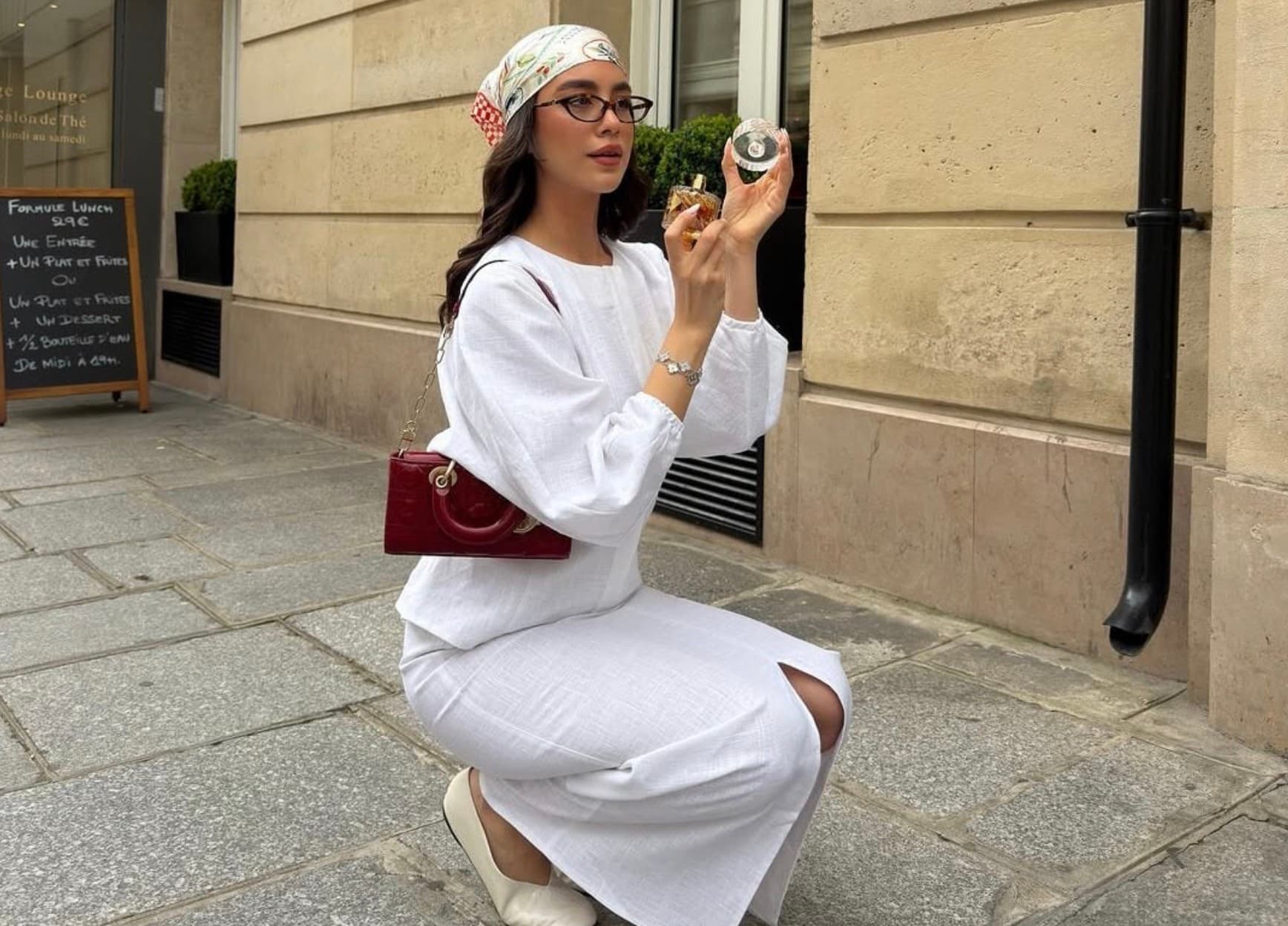 A woman in a white headscarf and glasses kneels on a Paris street holding a perfume bottle, wearing a red bag.